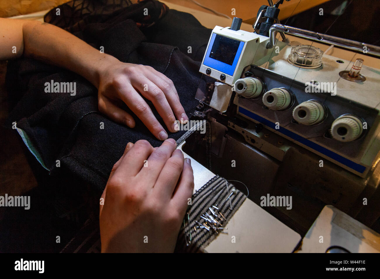 Mani di una sarta sono visti utilizzando una macchina taglia-e-cuci dentro la sua officina. Overlocking pezzi di tessuto durante lo sviluppo di vestiti. Foto Stock