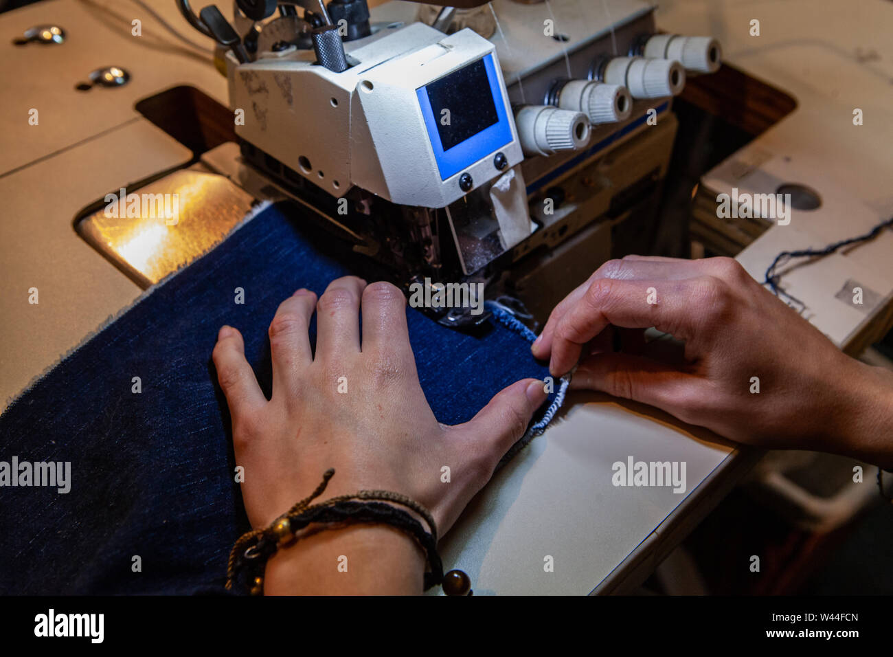 Un primo piano sulle mani di una sarta usando un industriale macchina overlocking per creare linee di giunzione in una chiazza di colore blu denim tessuto. Creazione di nuovo g Foto Stock