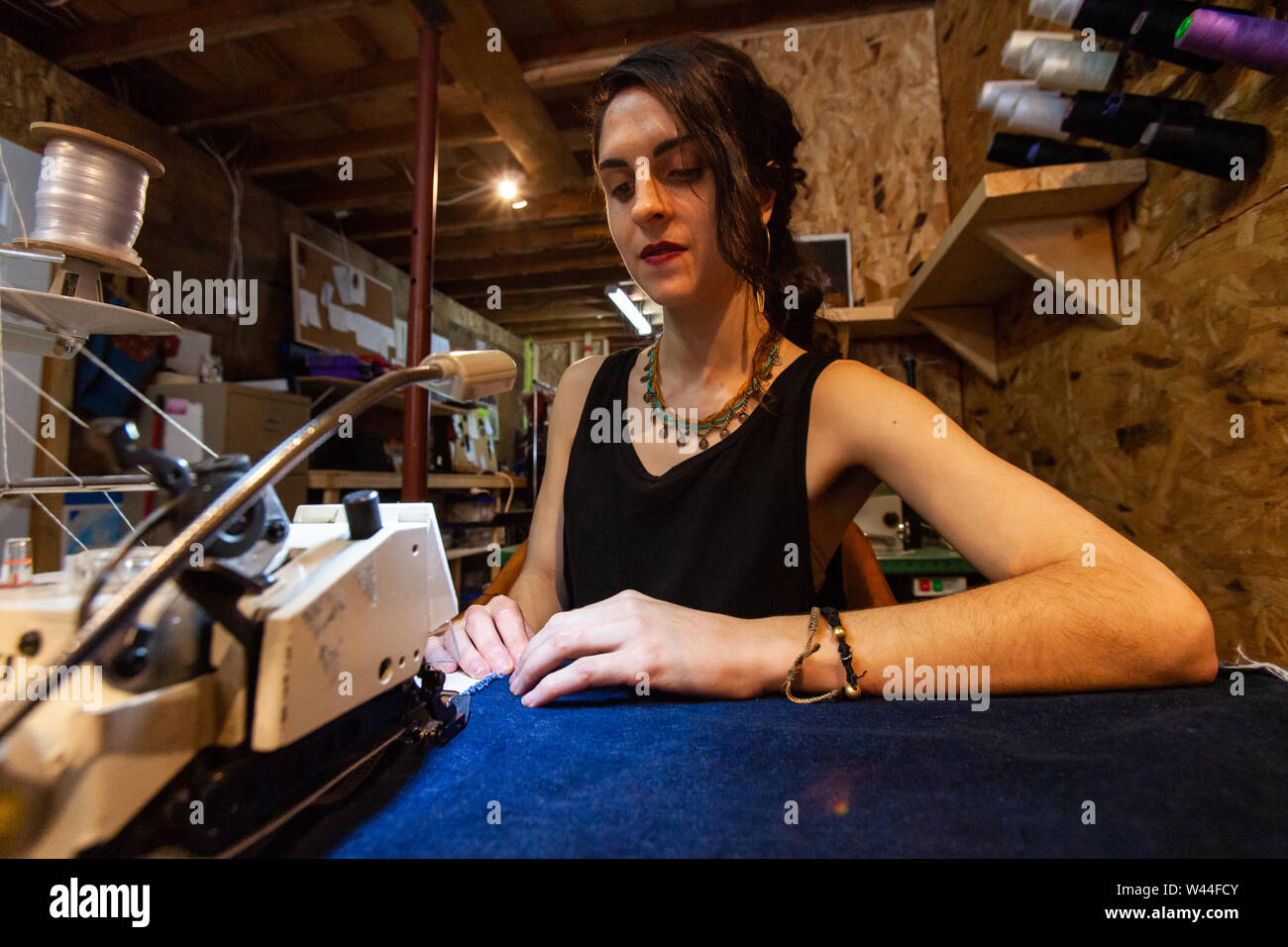 Una vista ravvicinata di una giovane sarta caucasica utilizzando una macchina di overlocking per creare una giunzione in tessuto blu, sat a workstation nel proprio studio creativo. Foto Stock