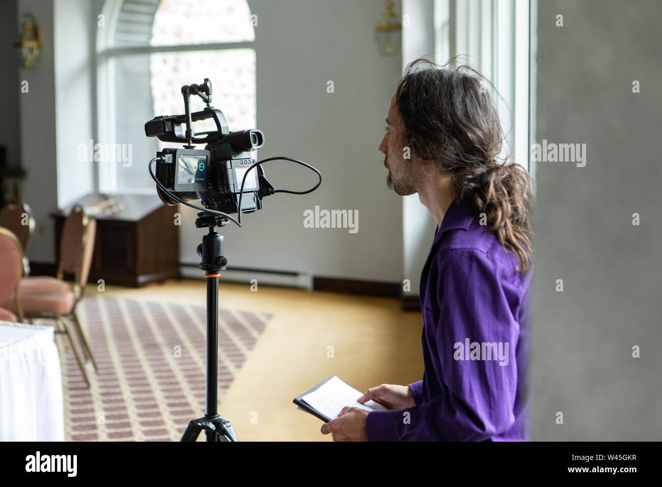 Un lato vista di profilo di un produttore cinematografico lavorando dietro una telecamera nel corso di una assemblea generale, le riprese di eventi e persone durante il giorno, con una camera Foto Stock