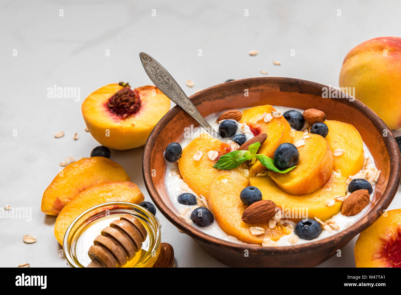 Yogurt con pesche, avena, mirtilli, miele e menta in una ciotola con un cucchiaio per una sana prima colazione su marmo bianco dello sfondo. close up Foto Stock