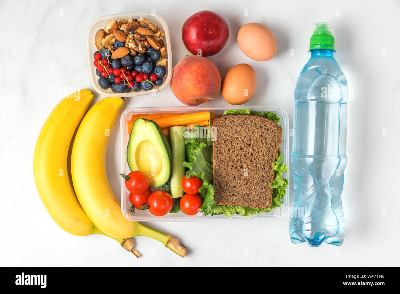 Pranzo a scuola di scatole con sandwich, verdure, dadi, bacche, banana, uova, pesche e acqua su marmo bianco dello sfondo. vista superiore Foto Stock