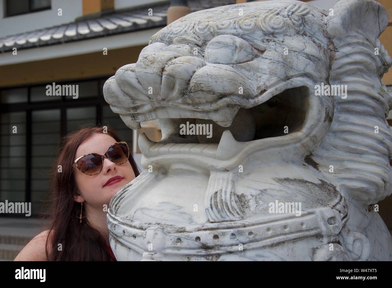 Un caucasico turista femminile con occhiali da sole pone con un grinning giapponese (asiatico) dragon (Lion) statua che si trova nella parte anteriore di un edificio Foto Stock