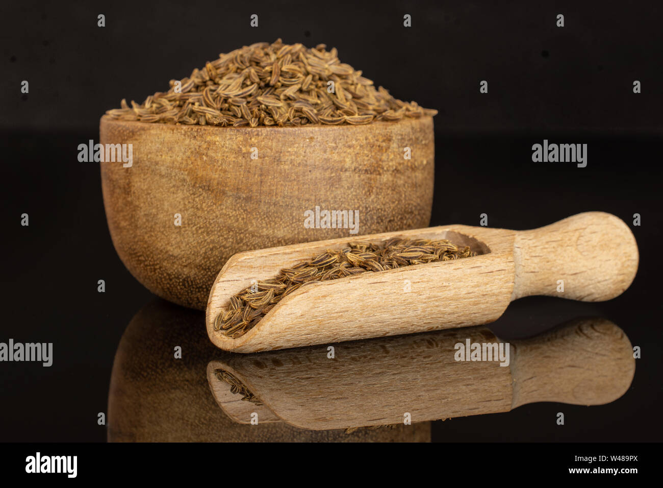 Sacco di tutta la secca di frutti di cumino in una ciotola di legno con il cucchiaio di legno isolata sul vetro nero Foto Stock