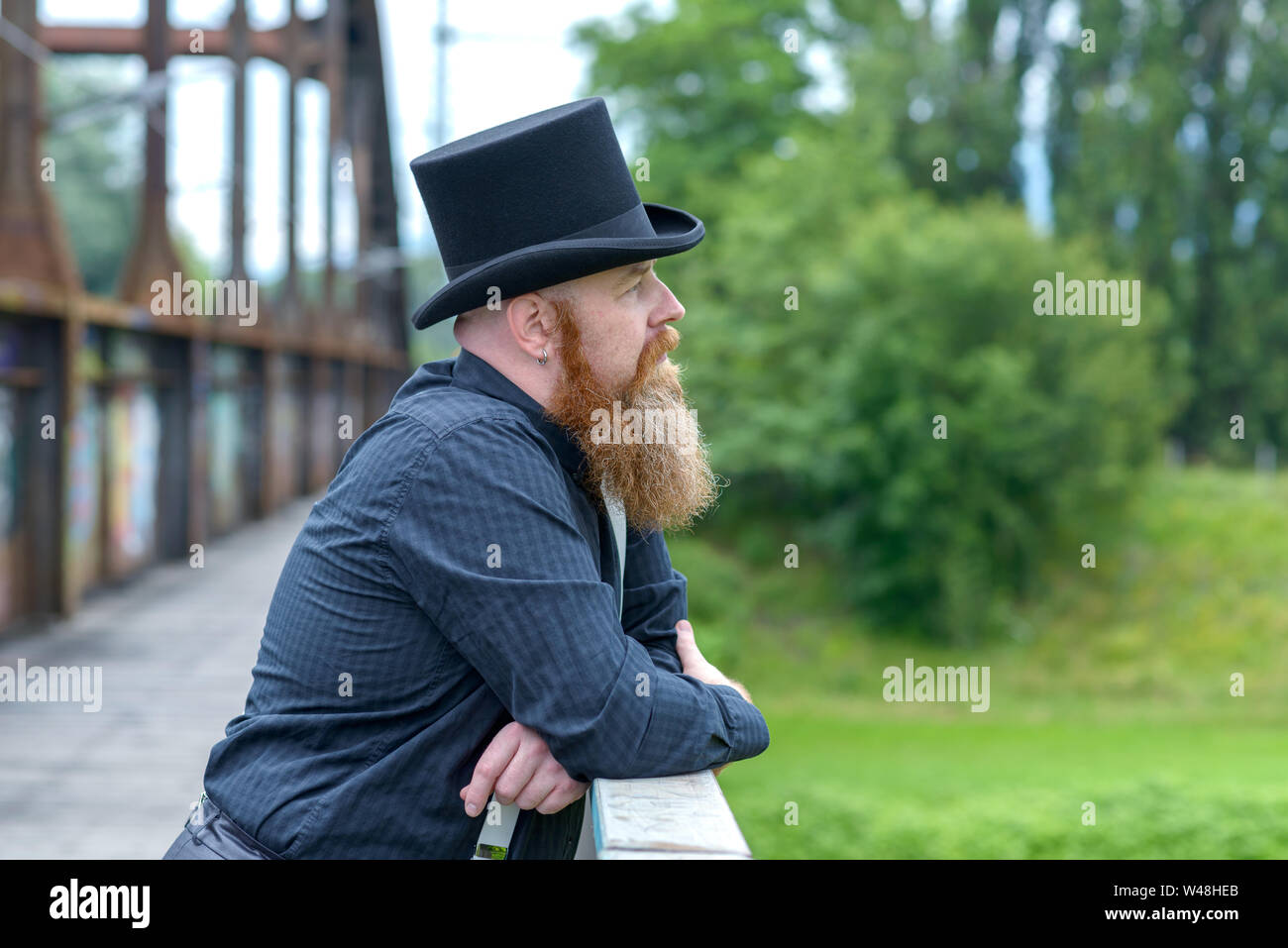 Uomo Barbuto in un top hat perso nel pensiero profondo come egli sta appoggiata sul parapetto di un ponte per la ricerca di Foto Stock