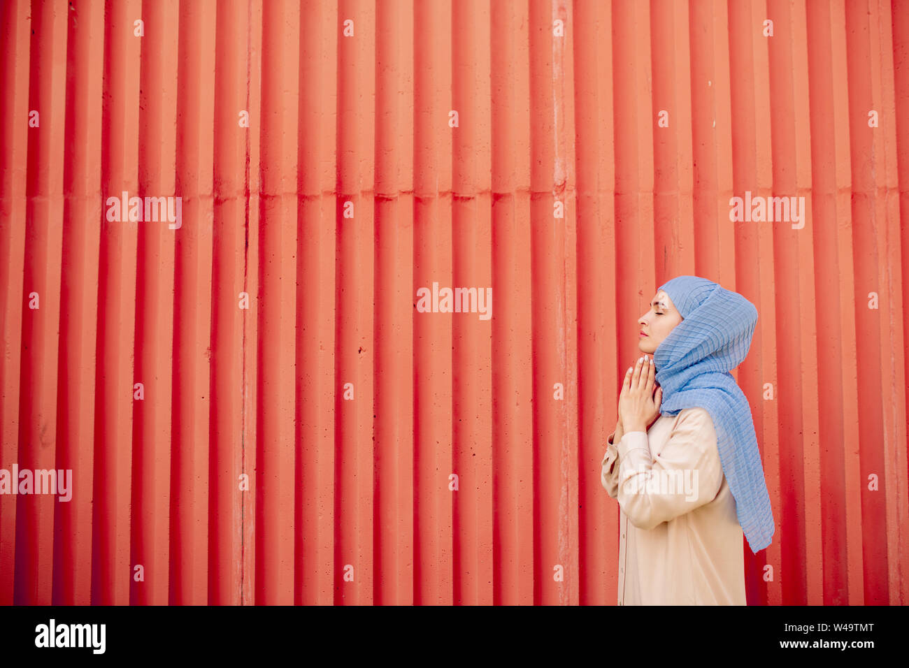 Giovane donna islamica in informale e hijab pregando contro la parete rossa Foto Stock