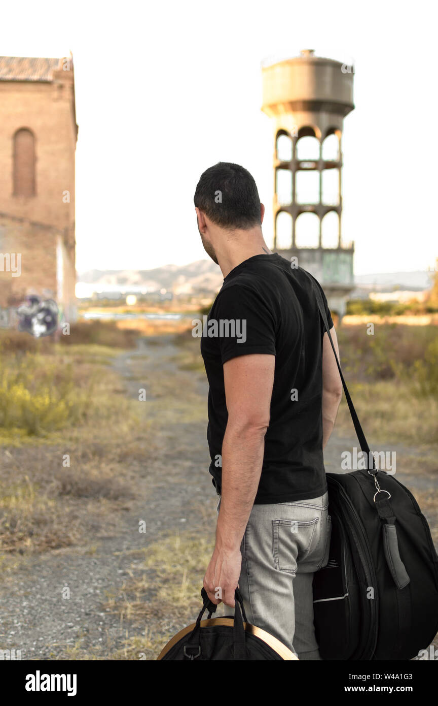 Uomo con abiti casual e bagagli persi nella periferia della città al tramonto. Esplorare la campagna e gli edifici abbandonati nelle periferie urbane. Foto Stock