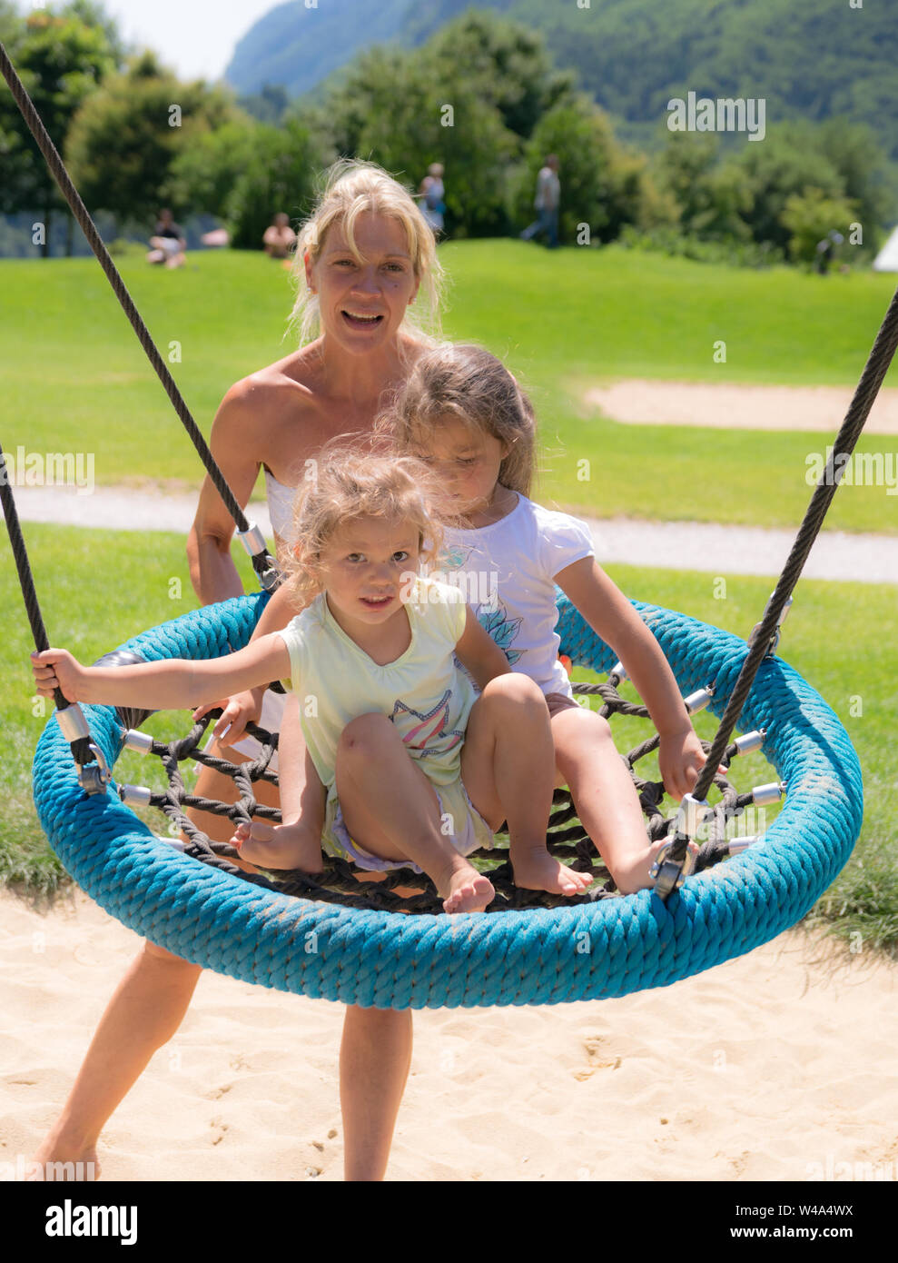 Molveno, luglio 10, 20151: Blonde mom spinge le ragazze sul round swing corda in un parco giochi. Foto Stock