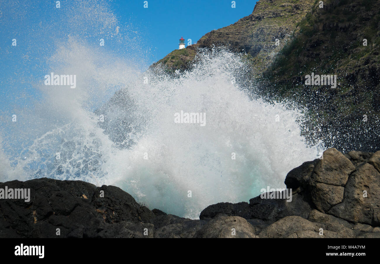 Vista di un onda di schiantarsi contro una costa rocciosa con il faro Mokapu in background. Questo è su Oahu, Hawaii's south shore. Foto Stock