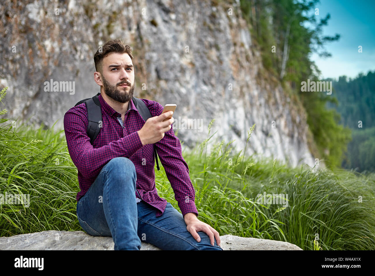 Un barbuto giovane uomo di ethnos caucasica possiede uno smartphone nelle sue mani, durante avventure su un sentiero escursionistico, riposa seduto su una pietra in ecozy Foto Stock