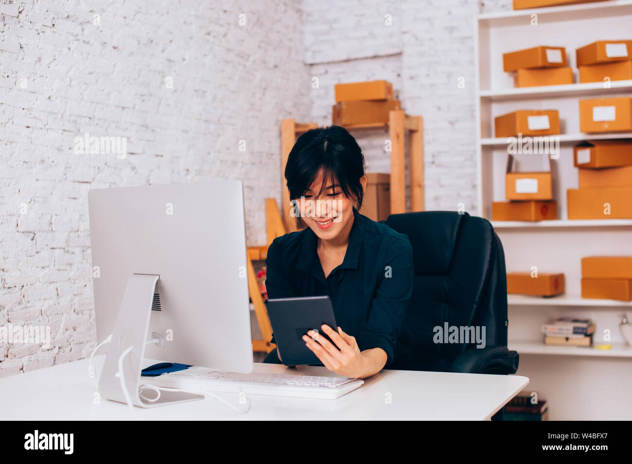 Allegro Asian imprenditrice sorridente e navigazione in internet tablet mentre è seduto alla scrivania con computer in ufficio Foto Stock