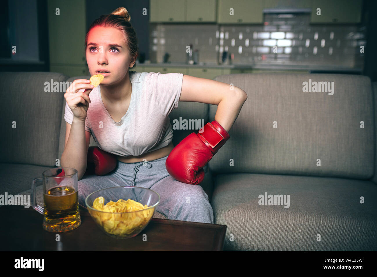 Giovane donna guarda il pugilato sulla tv di notte. Modello concentrato sporgersi in avanti e guardare in alto. Mangiare patatine. Il cibo spazzatura sul tavolo. Da soli in camera. Le tenebre. Guardare Foto Stock