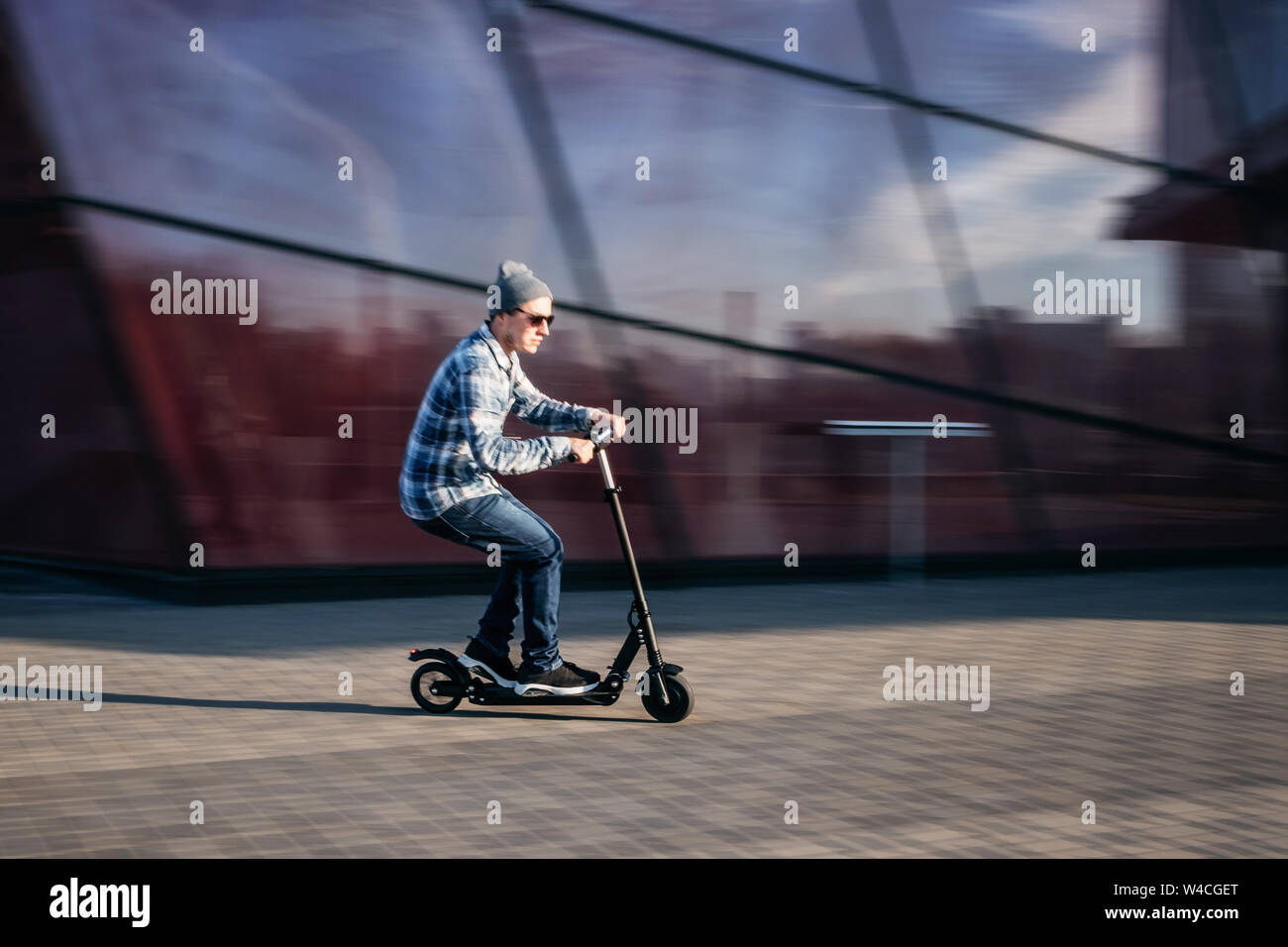 Giovane uomo in abbigliamento casual su elettrico kick scooter sulla strada di città in motion blur contro il vetro moderno edificio per uffici Foto Stock