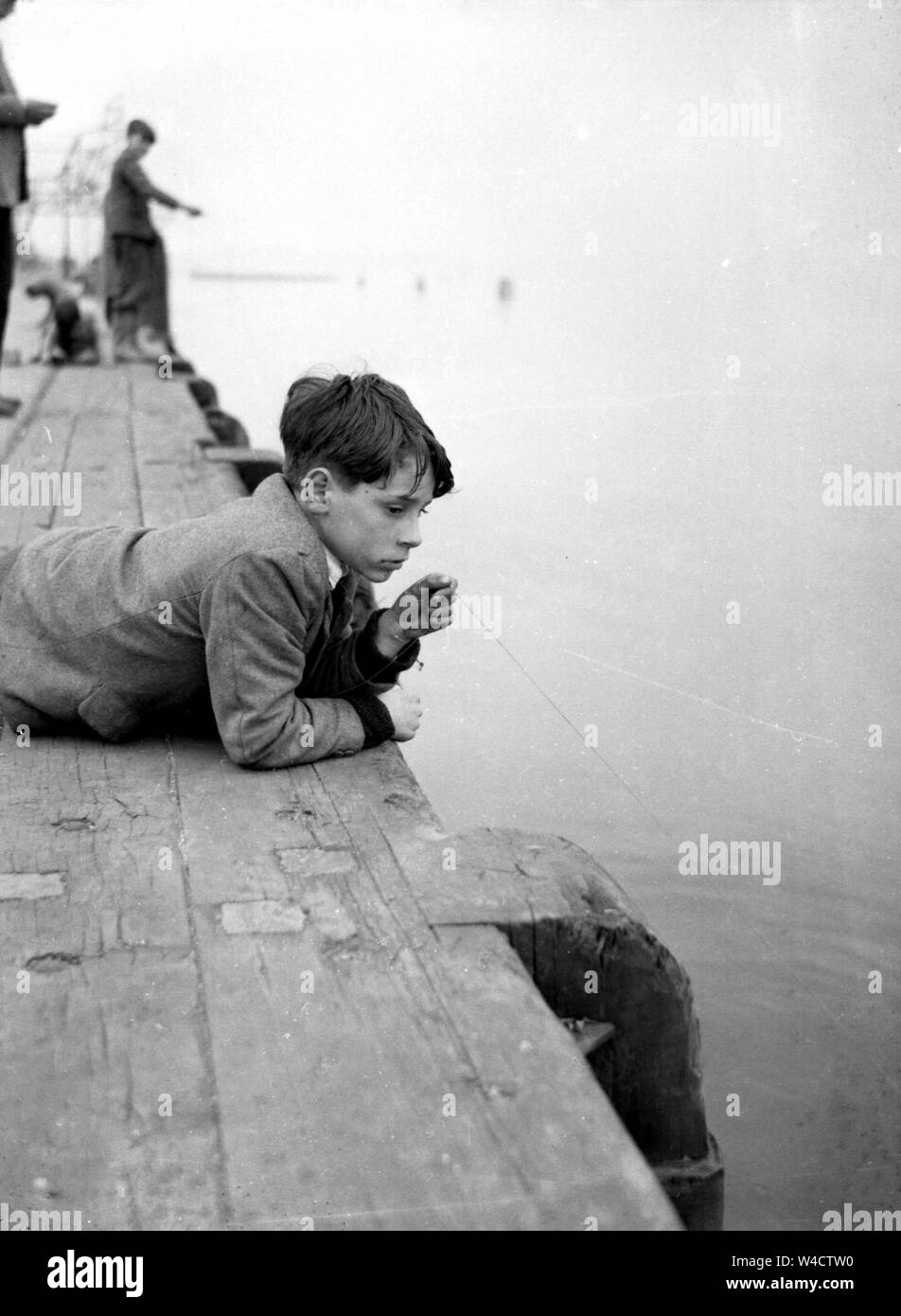 Un giovane ragazzo stabilisce a pancia in giù su un molo mentre la pesca con solo una linea negli anni trenta o quaranta Foto Stock