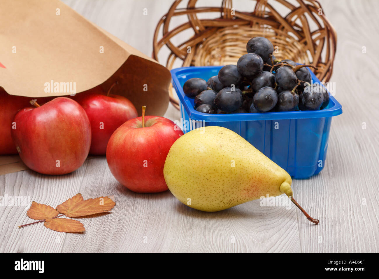 Giallo maturo pera, grappolo di uva in una scatola, un cesto di vimini, mele rosse con il sacchetto di carta e foglia secca sulla scrivania in legno. Un sano cibo biologico. Foto Stock