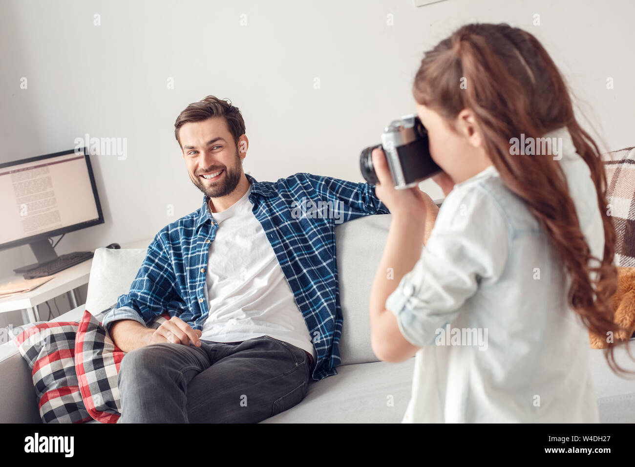 Padre e figlia piccola a casa ragazza tenere la fotocamera guardando indietro sorridente padre seduto sul divano allegro Foto Stock