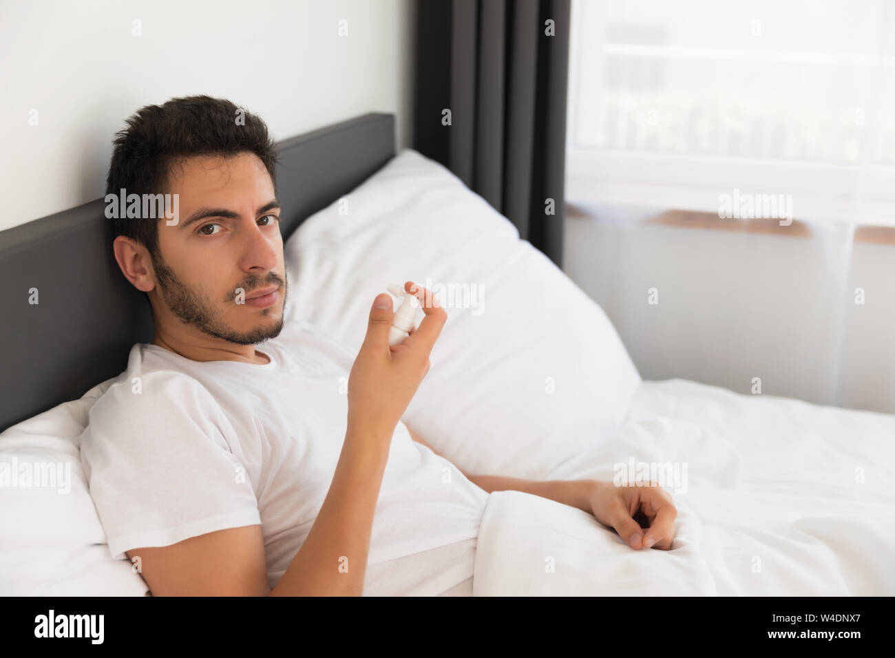 Un giovane uomo bello è seduto nel suo letto. Egli è in possesso di uno spruzzo con la medicina, egli ha bisogno di curare il mal di gola. Foto Stock