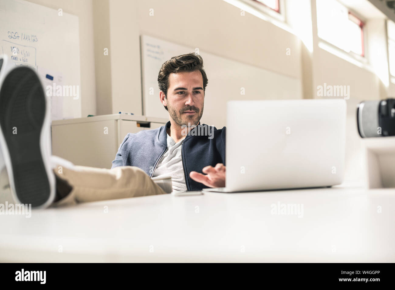 Giovane imprenditore lavora rilassato in un ufficio moderno, utilizzando laptop con piedi sulla scrivania Foto Stock