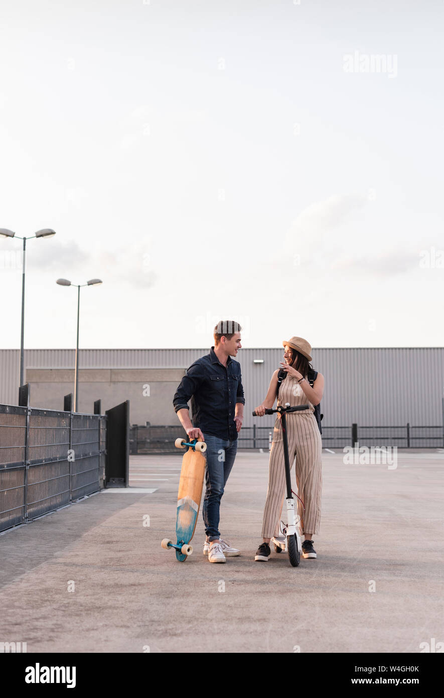 Giovane uomo e donna con longboard e scooter elettrici a parlare sul ponte di parcheggio Foto Stock