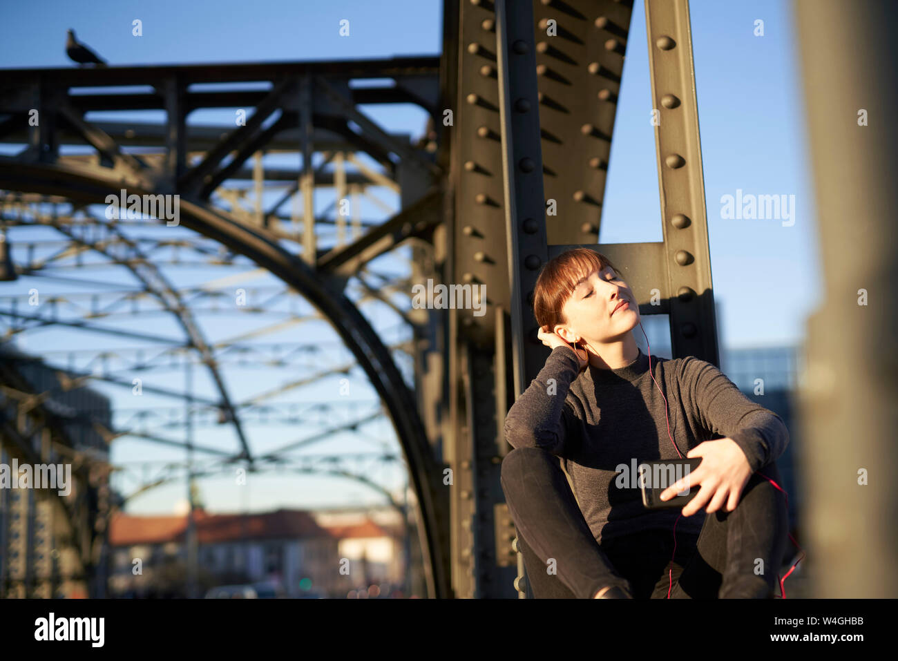 Ritratto di giovane rilassato donna seduta su trave in acciaio del ponte ascoltando musica con lo smartphone Foto Stock