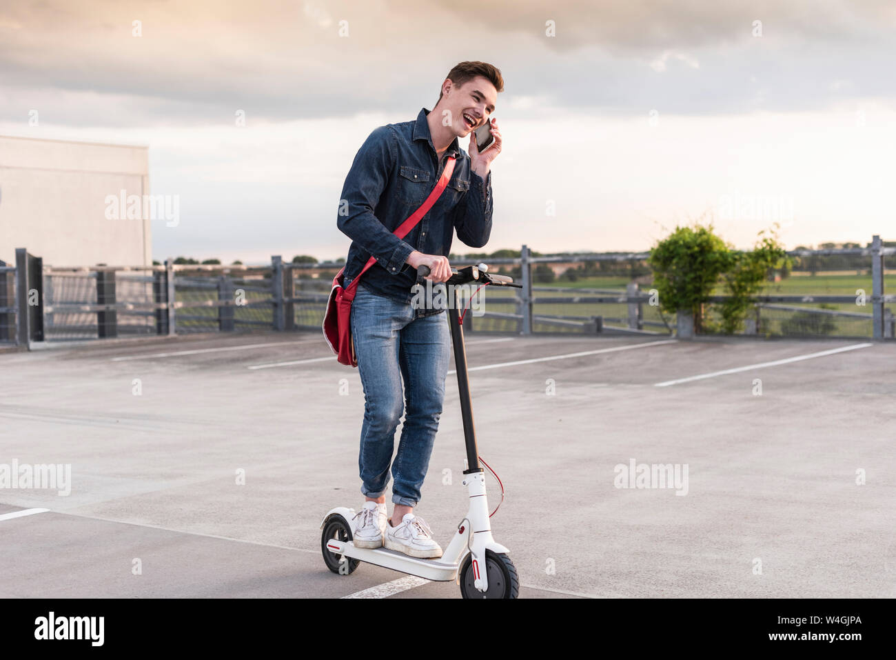 Felice giovane con un telefono cellulare e lo scooter elettrico sul ponte di parcheggio Foto Stock