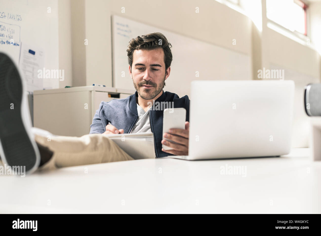 Giovane imprenditore lavora rilassato in un ufficio moderno utilizza lo smartphone e il computer portatile, Foto Stock
