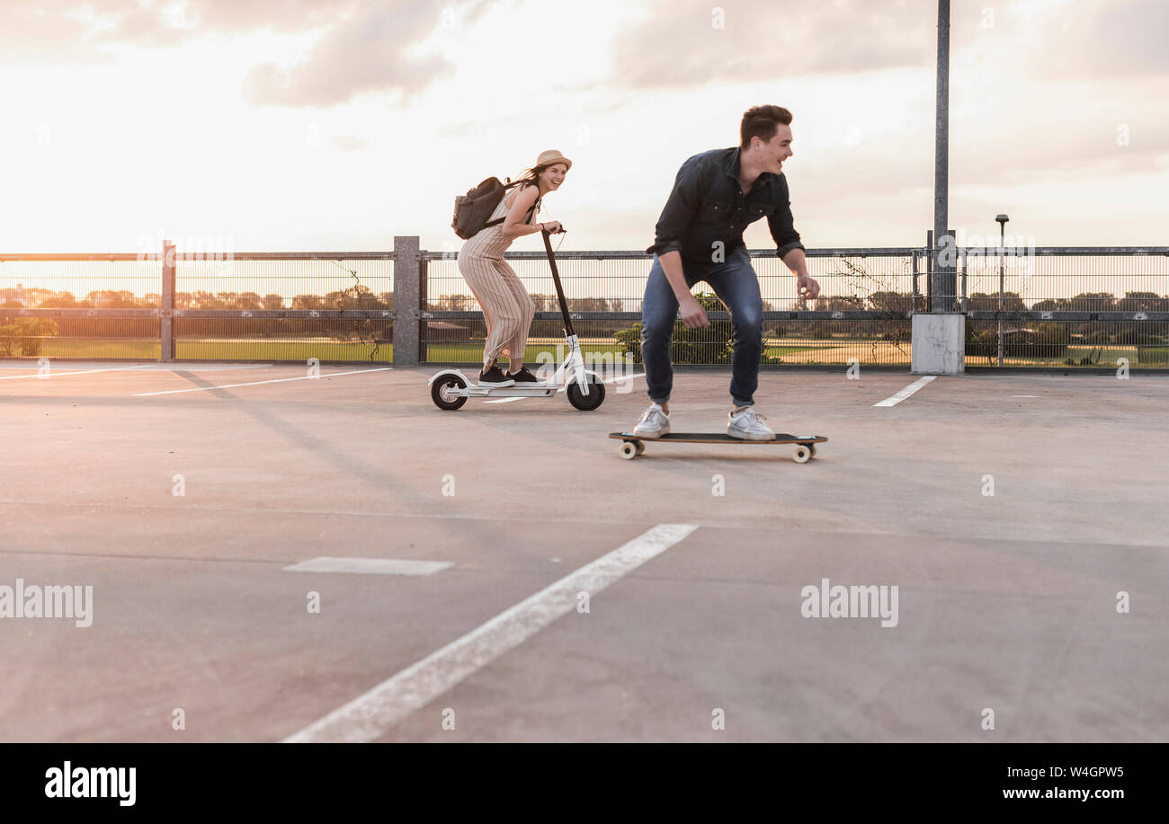 Giovane uomo e donna cavallo su longboard e scooter elettrico sul ponte di parcheggio al tramonto Foto Stock