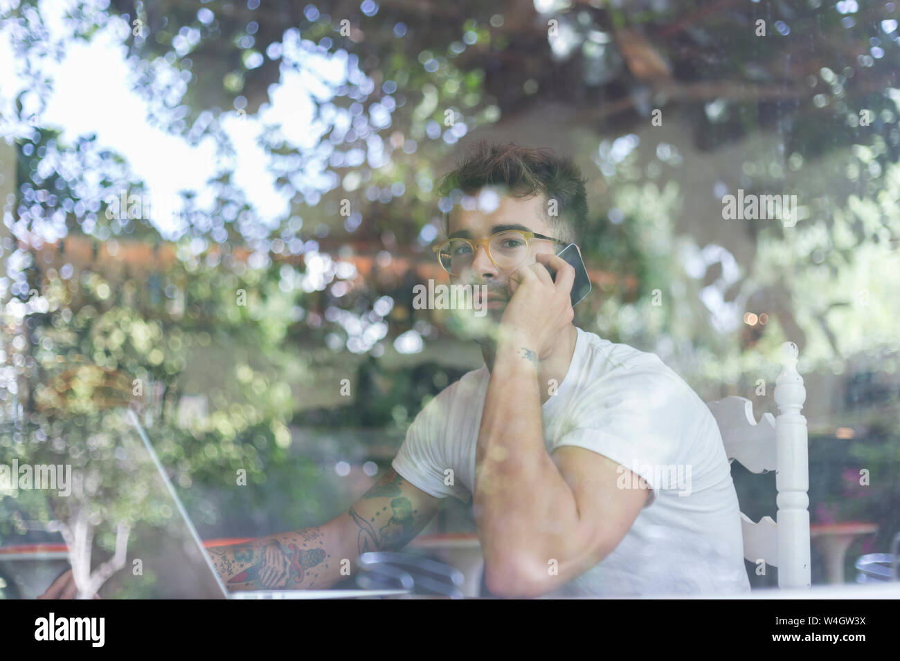 Giovane Uomo con laptop sul telefono cellulare dietro il vetro di finestra di un cafe Foto Stock