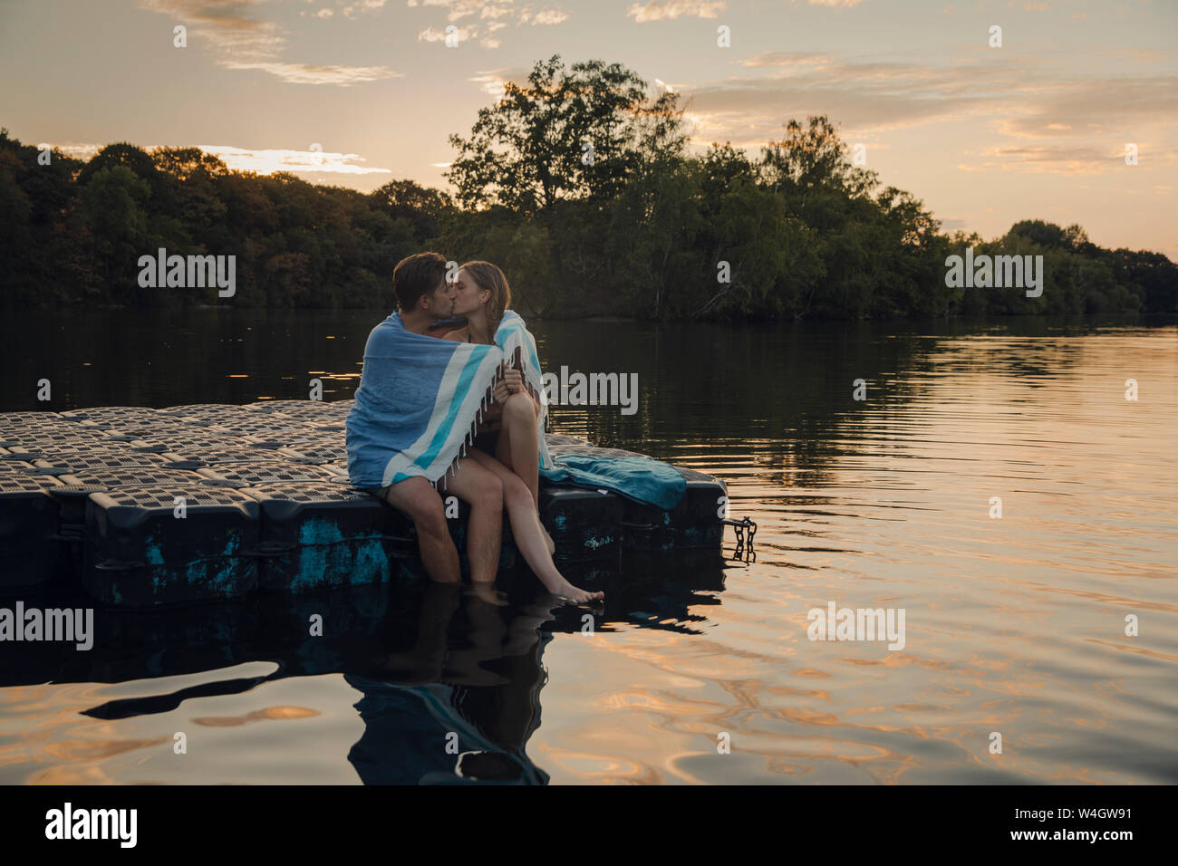 Coppia giovane seduto sulla piattaforma di balneazione, baciare, avvolto in asciugamano Foto Stock