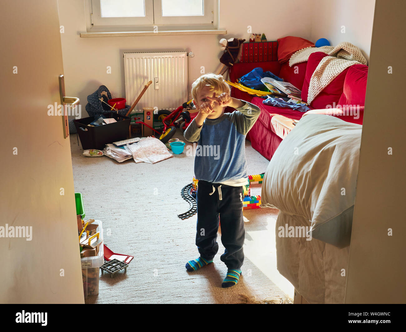 Little Boy in piedi nella sua camera disordinato edificio croce con le sue dita Foto Stock