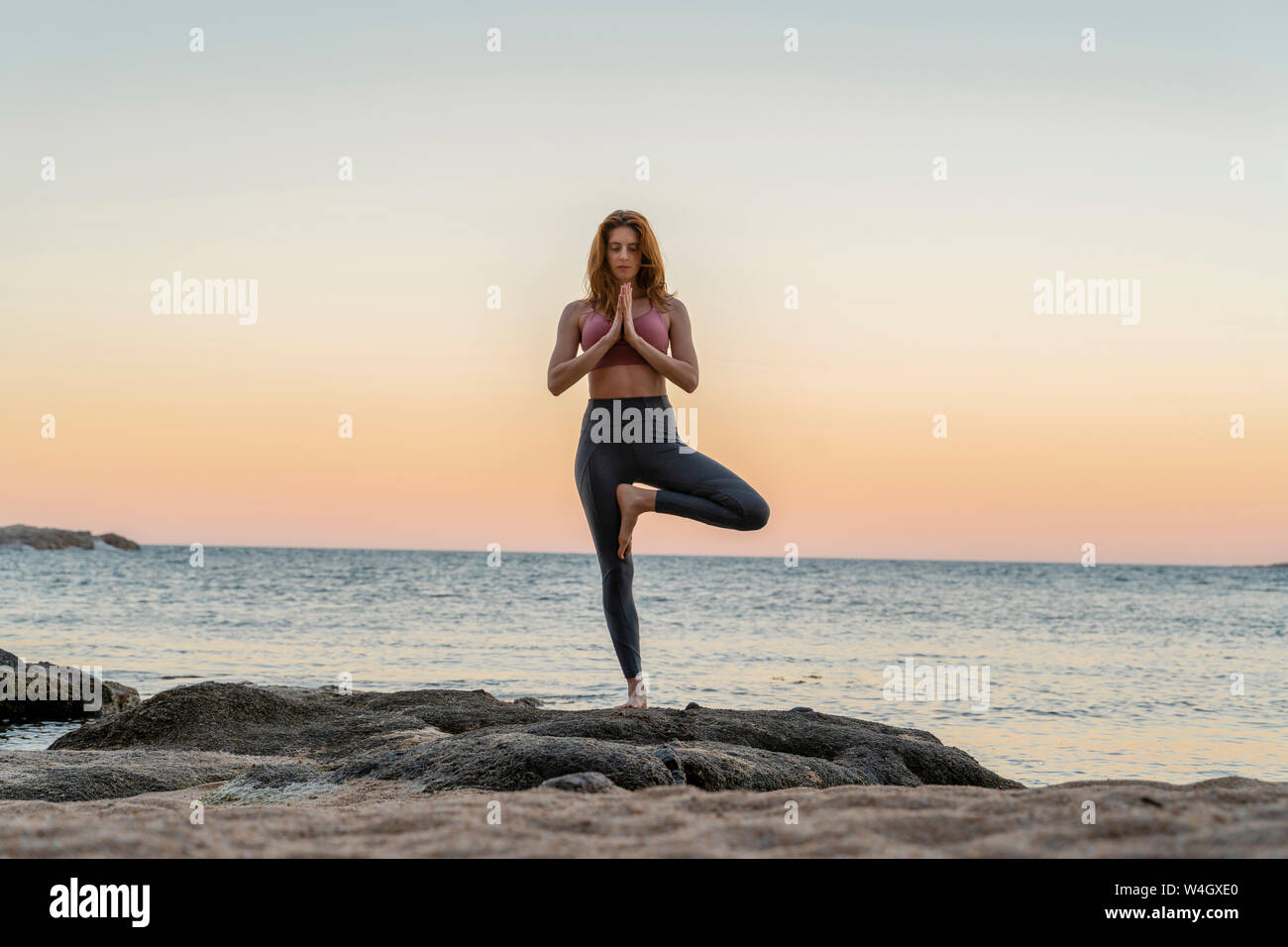 Giovane donna a praticare yoga sulla spiaggia, facendo la posizione dell'albero, durante il tramonto in spiaggia tranquilla, Costa Brava, Spagna Foto Stock