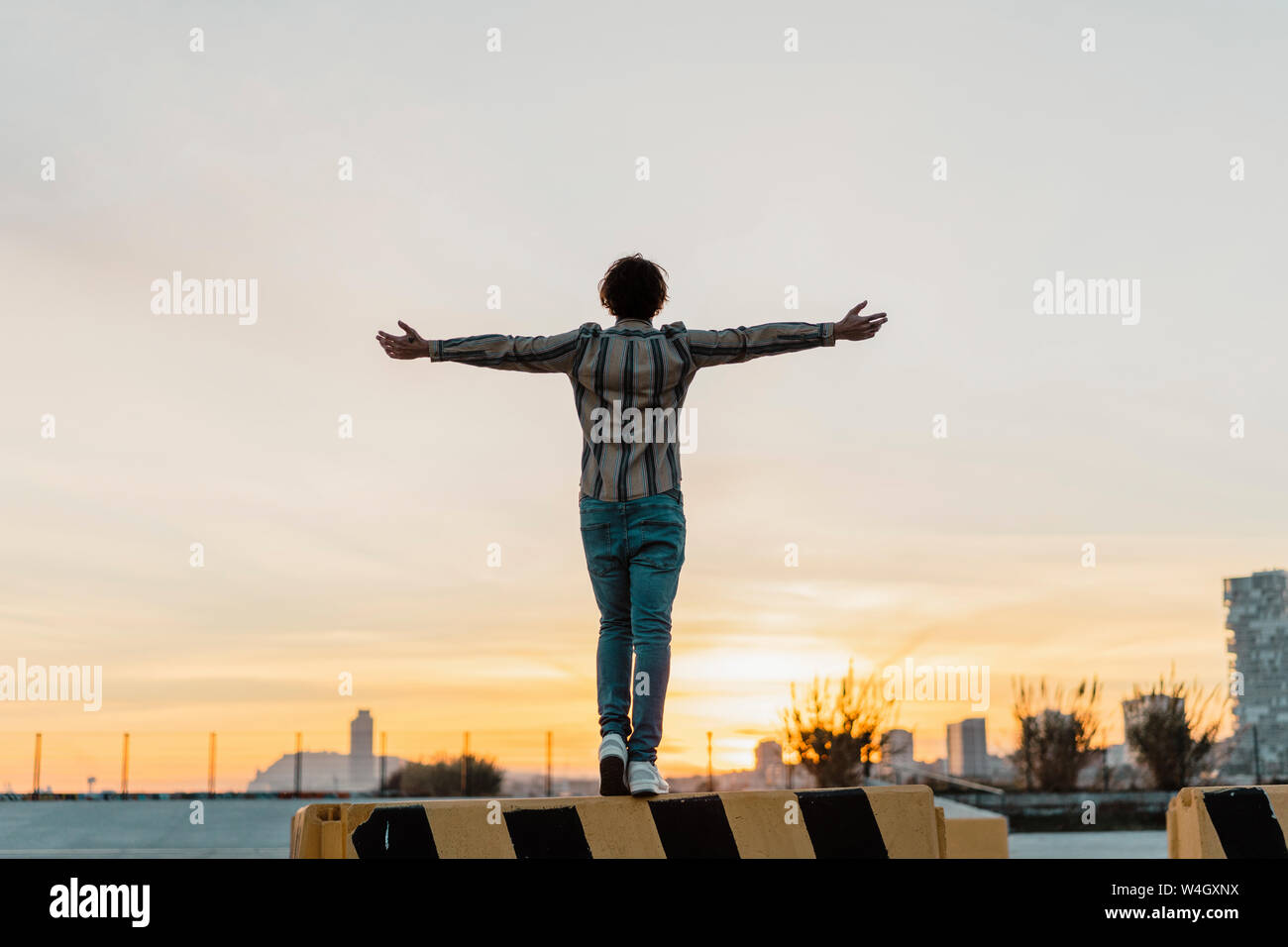 Vista posteriore dell'uomo con le braccia aperte in piedi sulla barriera godendo del tramonto, Barcellona, Spagna Foto Stock