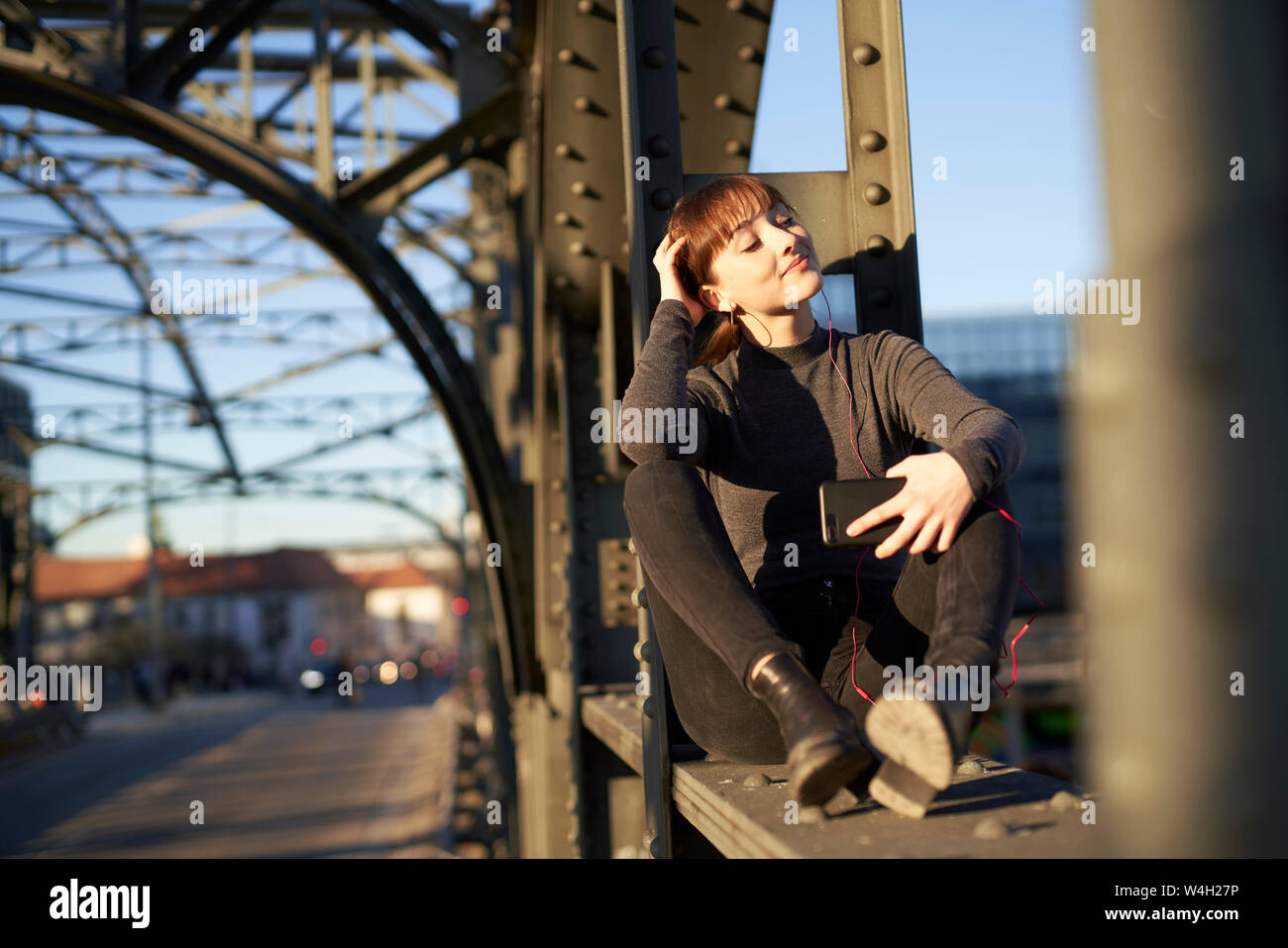 Ritratto di giovane rilassato donna seduta su trave in acciaio del ponte ascoltando musica con lo smartphone Foto Stock