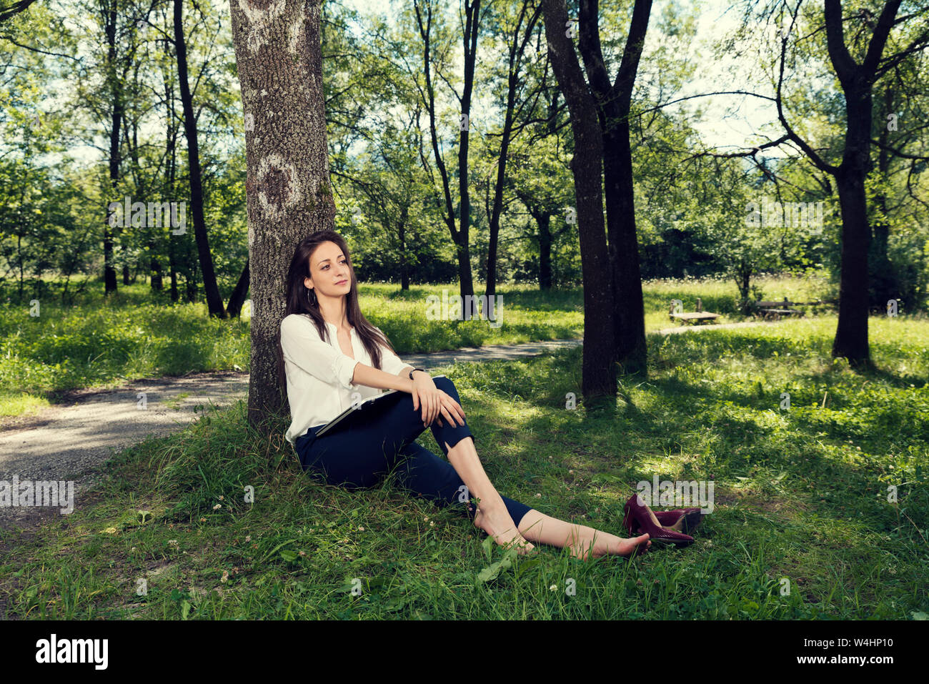 Business donna seduta di appoggio sotto l'albero senza scarpe in un pubblico parco della città messa a fuoco selettiva Foto Stock