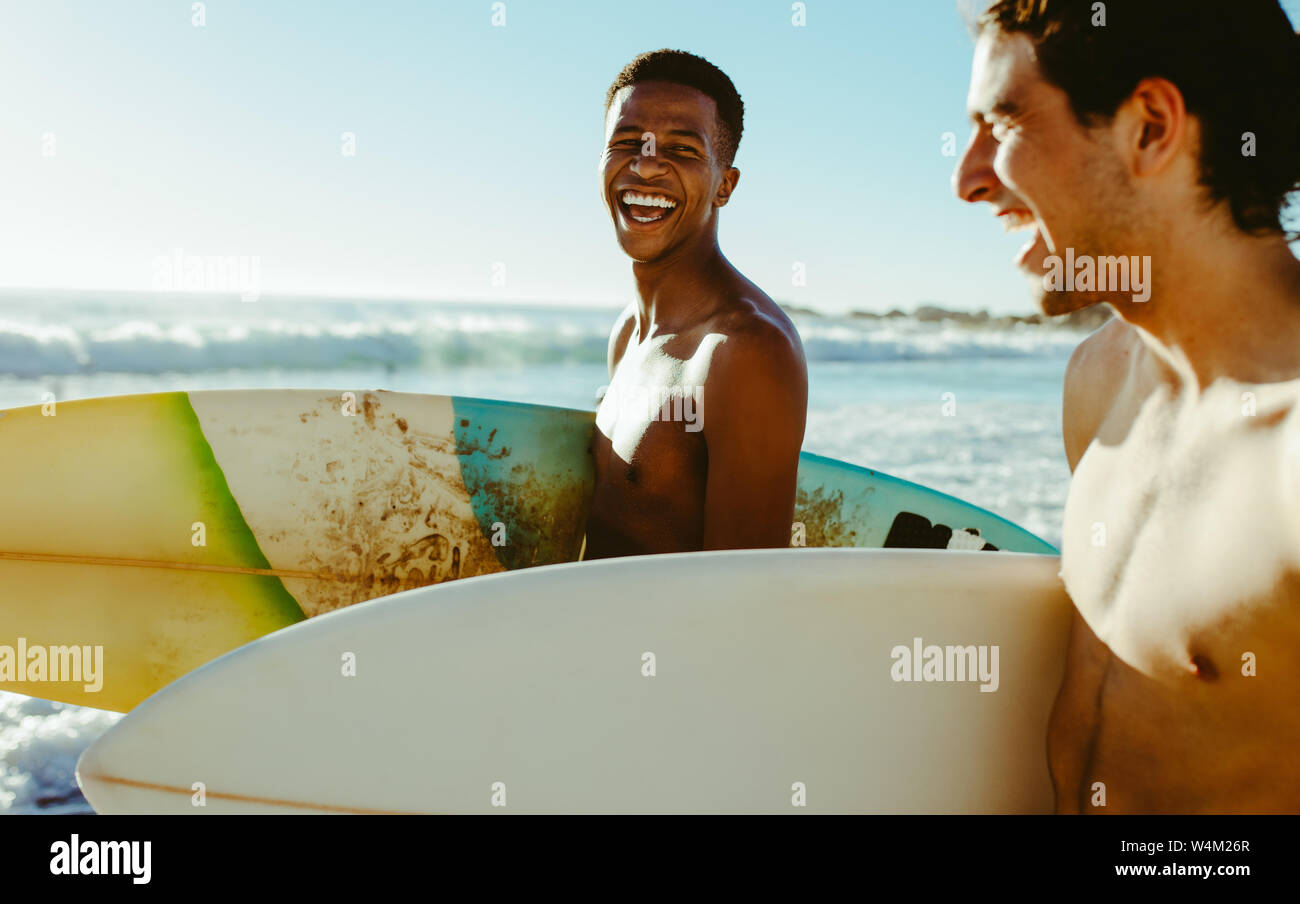 Felice giovane con un amico di camminare sulla spiaggia che trasportano le tavole da surf. Allegro giovani amici in vacanza al mare. Foto Stock