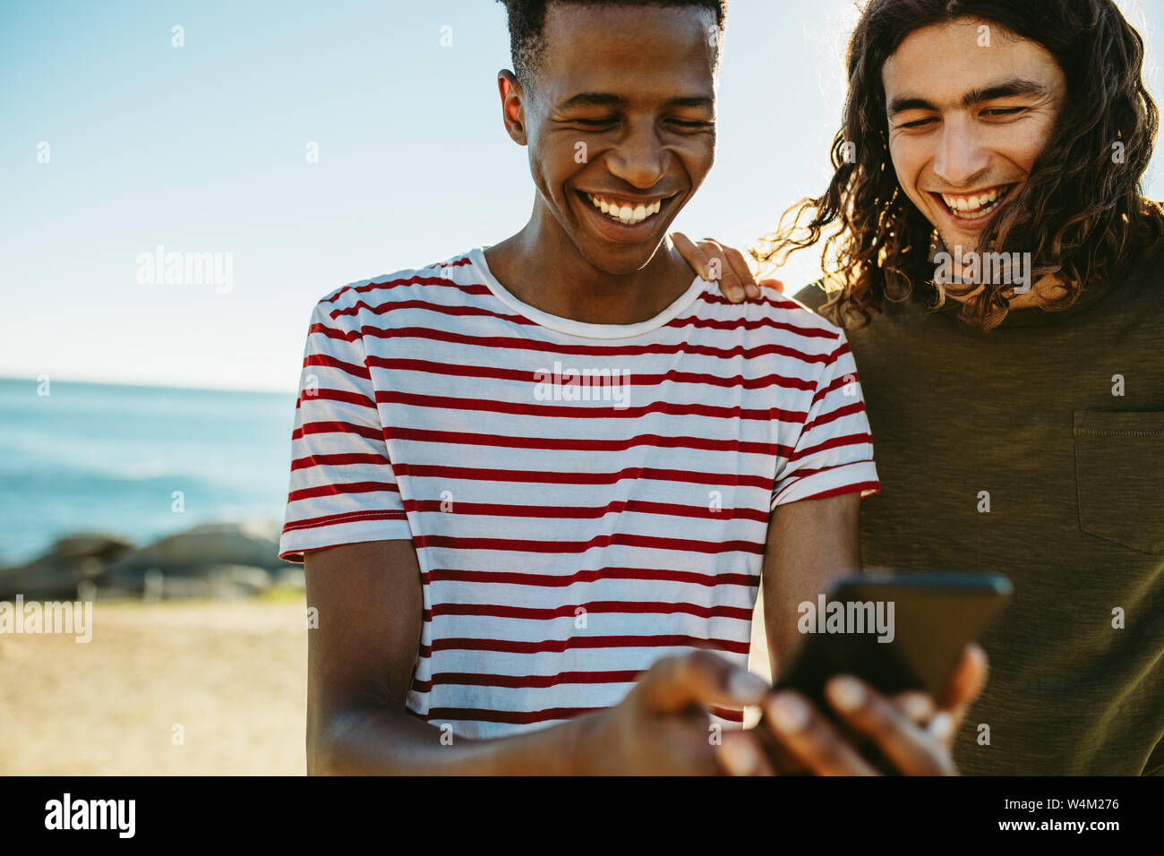 Due giovani amici all'esterno guardando al telefono cellulare e sorridente. Giovani uomini utilizzando uno smart phone dalla spiaggia. Foto Stock