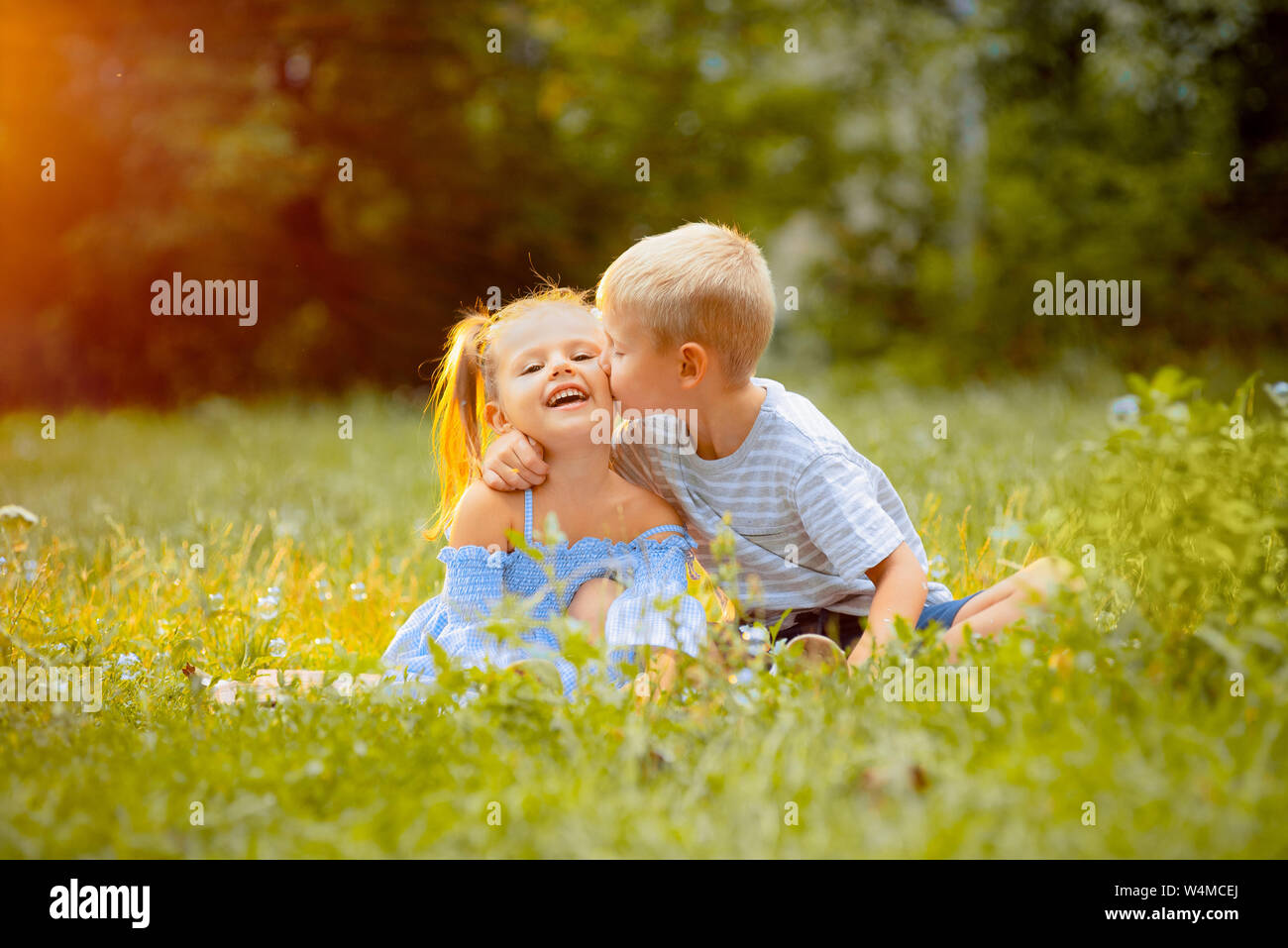 Bellissimi bambini di sedersi su un prato verde in raggi di un tramonto. Il ragazzo baci e abbracci la ragazza sulla guancia. Dei bambini felici Foto Stock