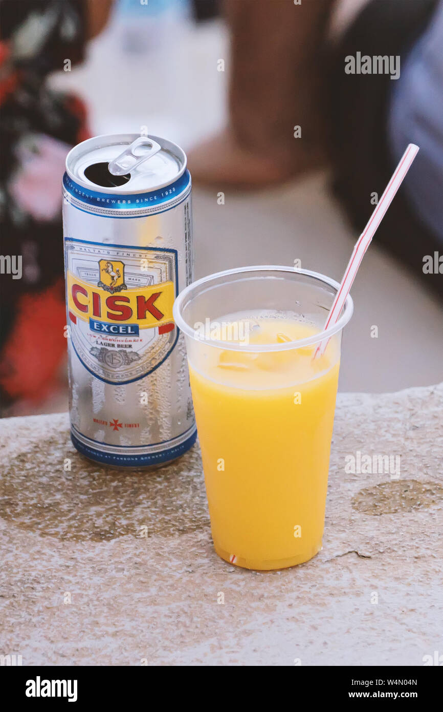 La Valletta, Malta - 28 Giugno 2019: Può di maltese Cisk locale birra lager e il succo di arancia in una tazza di plastica con paglia sulla strada Foto Stock