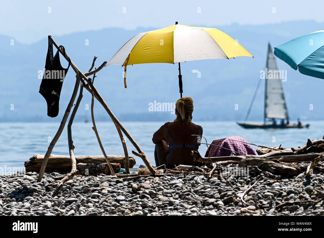 24 luglio 2019, Baden-Wuerttemberg, Gohren am Bodensee: una donna siede sulla riva del lago di Costanza sotto un ombrellone mentre un vele di imbarcazioni in background. Foto: Felix Kästle/dpa Foto Stock