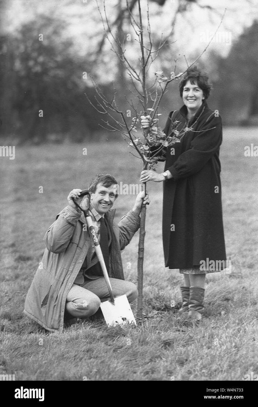 Anni '1980, storico, 'tress are good'....un uomo e una donna fuori in un campo in piedi accanto a un albero appena piantato, noto come alberello, Inghilterra, Regno Unito. Stanno proponendo un quadro pubblicitario per evidenziare l'importanza degli alberi e il contributo che essi apportano per invertire l'impatto del cambiamento climatico e migliorare l'ambiente condiviso per le comunità. Foto Stock