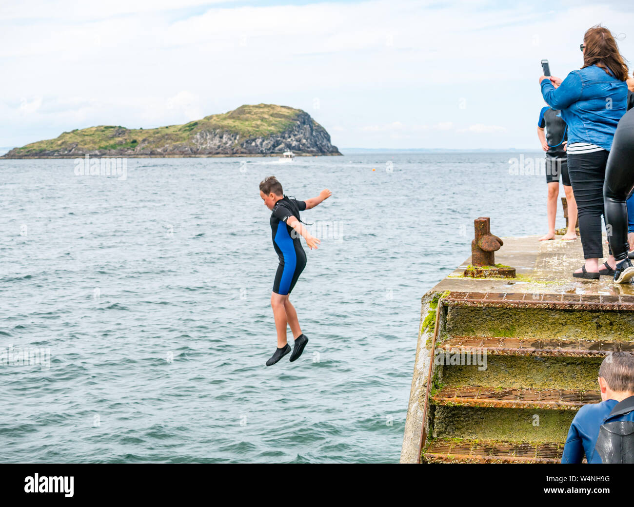 A North Berwick, East Lothian, Scozia, Regno Unito, 24 luglio 2019. Regno Unito Meteo:un ragazzo che indossa una muta copes con la canicola estiva che salta fuori il vecchio molo nel Firth of Forth per rinfrescarsi con Craigleith Island in background Foto Stock