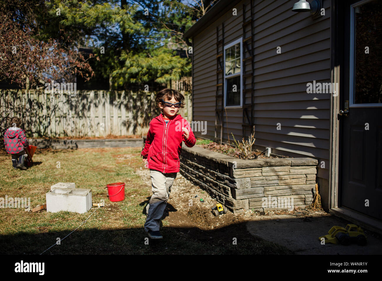 Un bambino piccolo in una felpa rossa e occhiali da sole corre nel cortile soleggiato Foto Stock