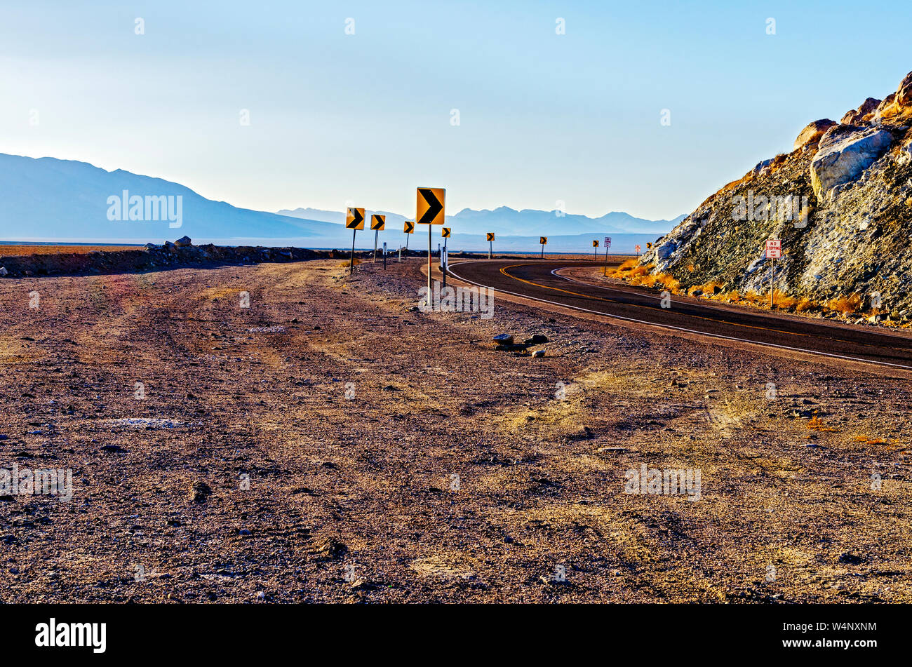 Autostrada deserta curvando attorno al colle roccioso con sopito delle montagne del deserto al di là. Freccia che indica i segni della curvatura road. Foto Stock