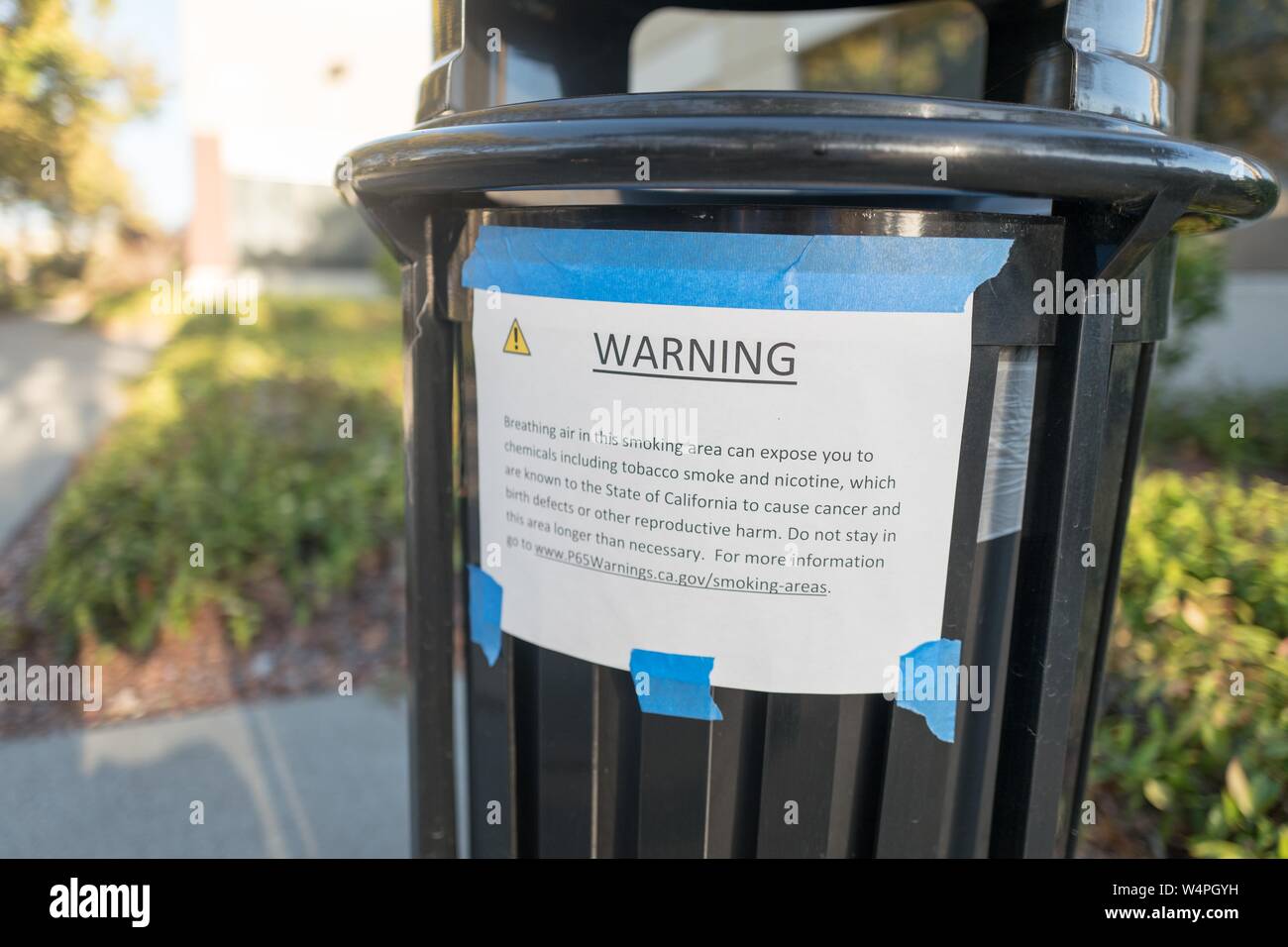 Proposta stampato 65 avviso apposto sul lato di un outdoor trash can in un ufficio parco sulla isola di Alameda, Alameda, California, avvertimento che la zona può essere contenuta con il fumo di tabacco a causa della presenza di una area fumatori, 10 settembre 2018. () Foto Stock