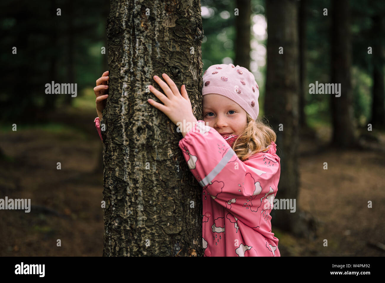 Bimba bionda indossando un impermeabile rosa e hat abbracciando albero a Reykjavik, Islanda foresta guardando la fotocamera Foto Stock