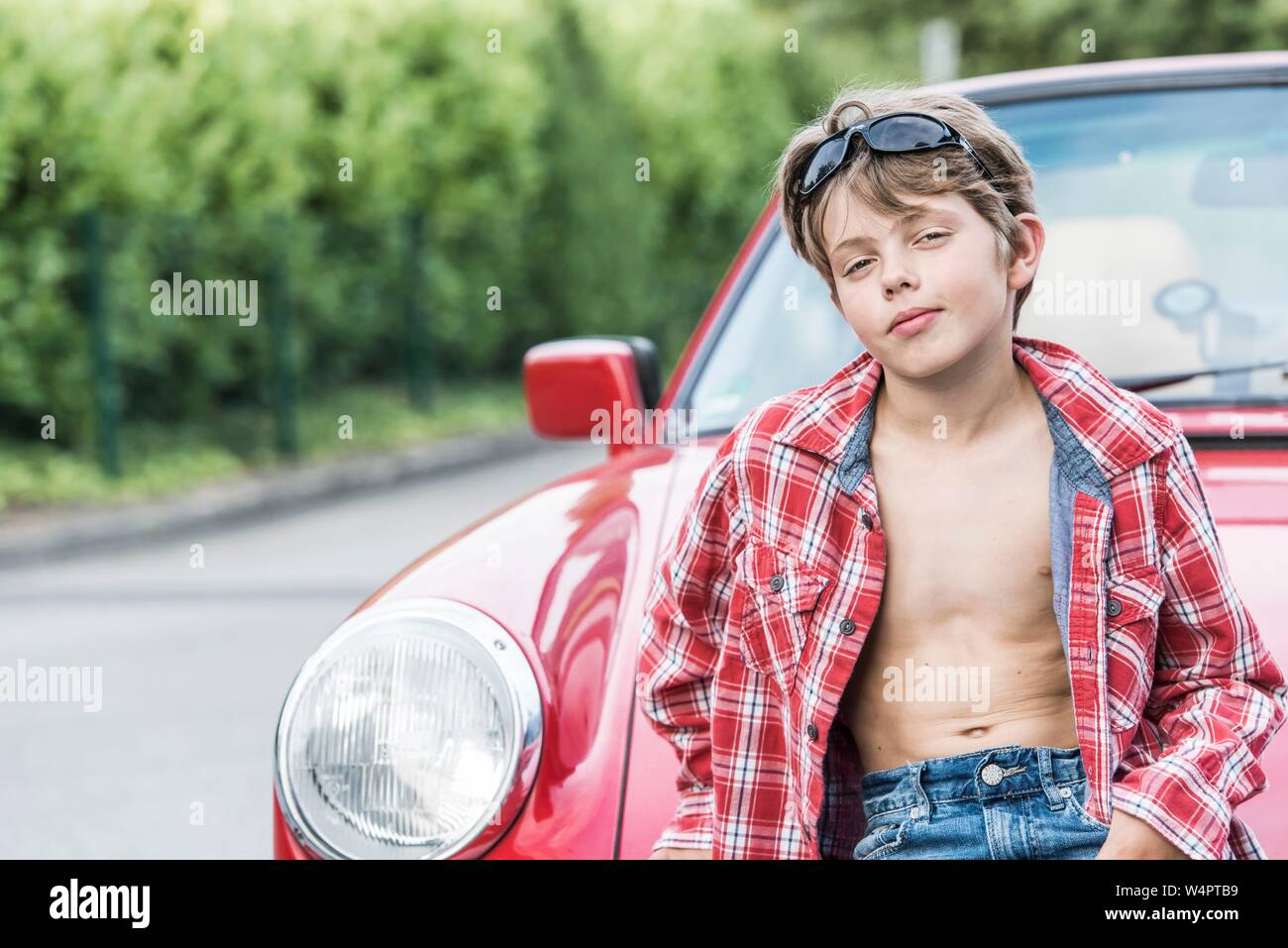Ragazzo, dieci anni, con occhiali da sole e plaid shirt poggia su una automobile rossa e guarda a raffreddare nella fotocamera, Germania Foto Stock
