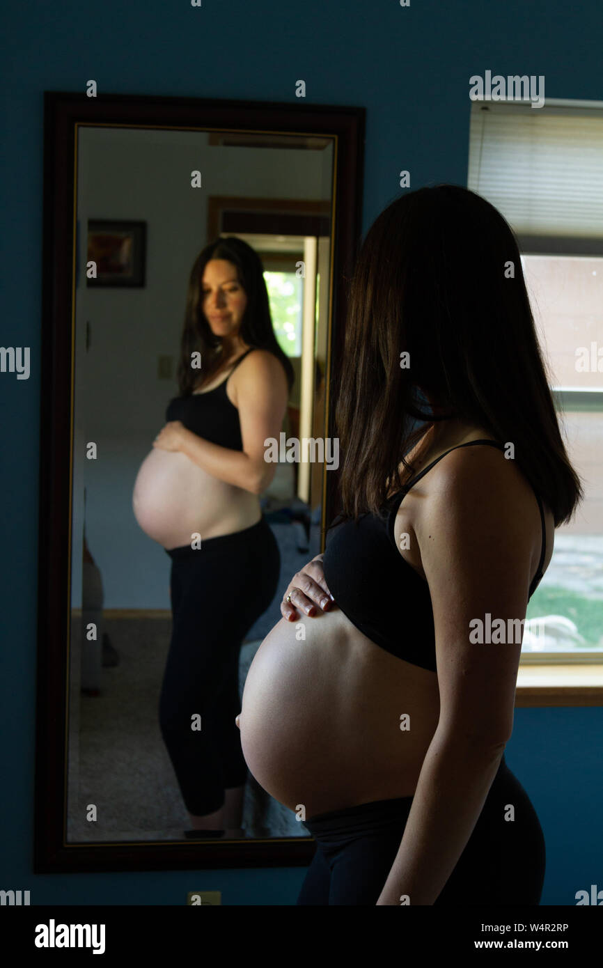 Donna incinta tardi nel terzo trimestre cercando longingly al suo bambino bump nello specchio. Ella è altamente anticipando l'arrivo del bambino. Foto Stock