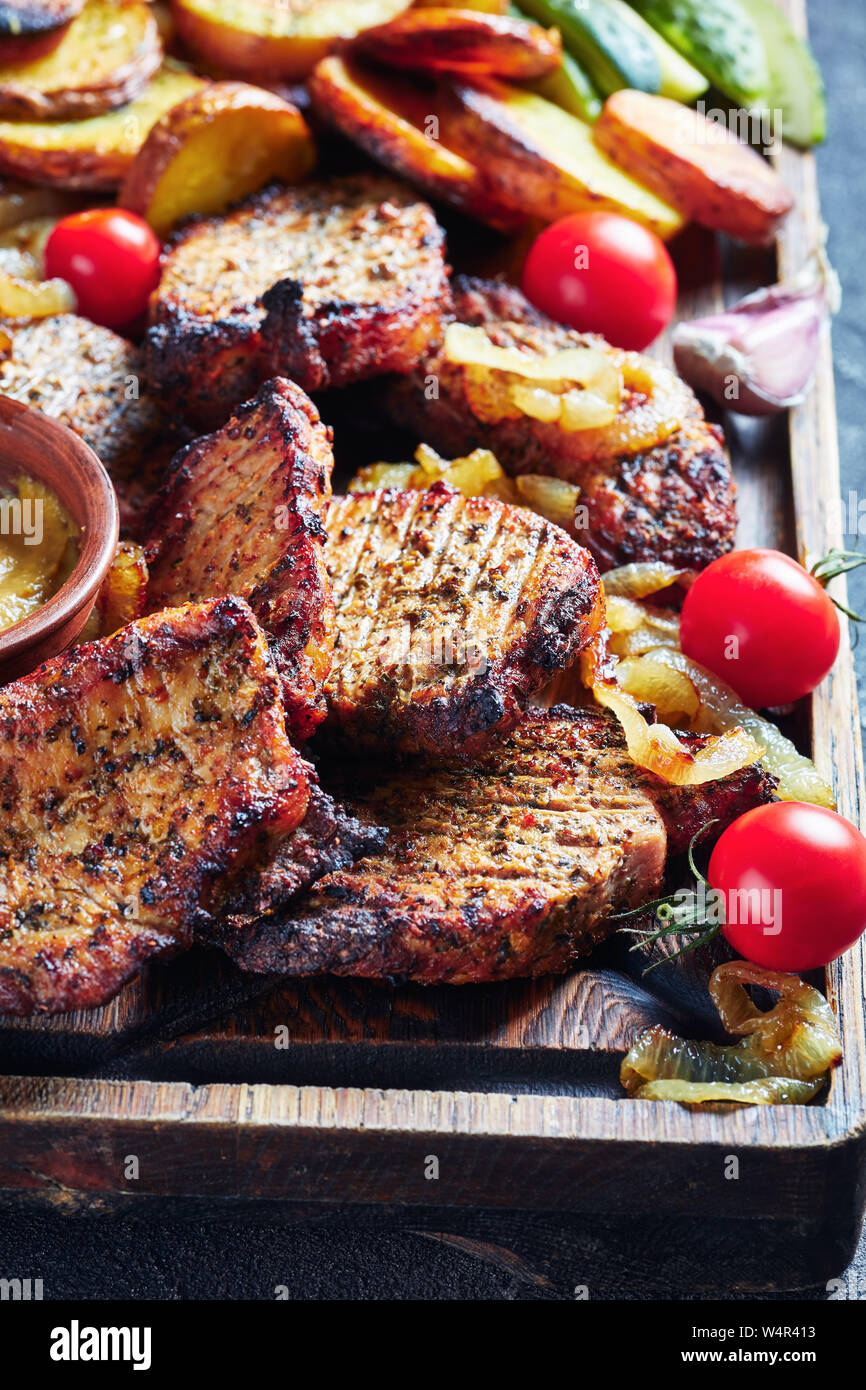 Close-up di Schwenkbraten, succosa, fumoso tedesco alla griglia costolette di maiale servito su un tagliere con fette di patate fritte, cipolle, pomodori e cetrioli, Foto Stock