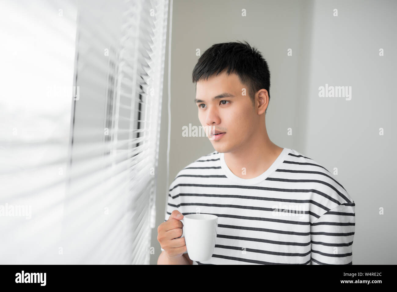 Giovane bello spensierato uomo moderno vicino a finestre di lunghezza completa gustando una tazza di caffè mentre guardando al di fuori, di buon mattino, Foto Stock