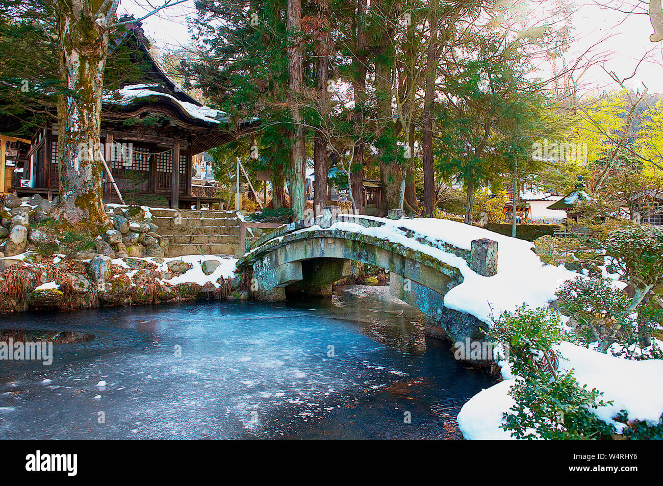 Passerella di pietra ricoperta di neve oltre il fiume che porta alla piccola casa in legno, Hida-Takayama, Takayama, Prefettura di Gifu, Giappone Foto Stock